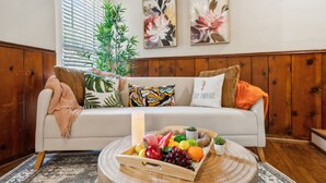 A cozy living room with a wooden panel wall, a white sofa with colorful cushions, a round wooden coffee table with a fruit tray, and a potted plant. - This is Your Home Away From Home! Warm & Comfy 2BD (Fort Worth)
