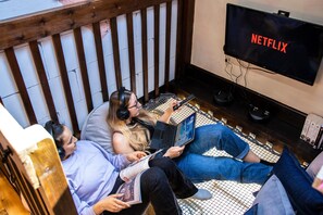 Ein Zimmer mit Holzboden und einer Decke aus Holzleisten, einem Fernseher mit dem Schriftzug 'NETFLIX' und einer Person, die einen Laptop benutzt.