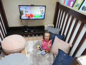Ein Kind sitzt auf einem grauen, flauschigen Teppich in einem Raum mit einem Fernseher, der eine Zeichentricksendung zeigt, einem Holzstuhl und einem Bücherregal mit Büchern.