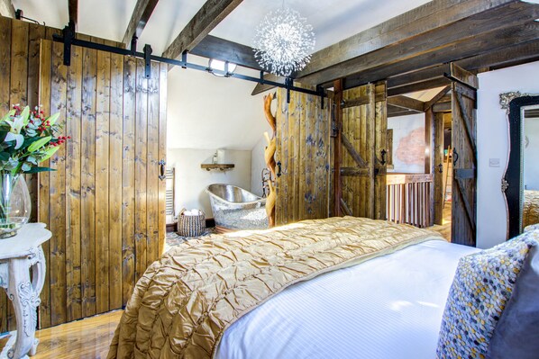 A rustic bedroom with a wooden slat wall, a bed with a beige quilt, a white lamp, and a large mirror.