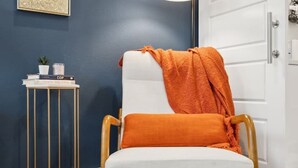 A chair with a white cushion and an orange throw blanket, a floor lamp, a small white side table with a plant and books, and a blue wall with a framed artwork. - The Charm :2BD In the Heart of Magnolia & Stylish! (Fort Worth)