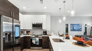 A modern kitchen with stainless steel appliances, a central island, and a dining area with orange chairs. - The Charm :2BD In the Heart of Magnolia & Stylish! (Fort Worth)