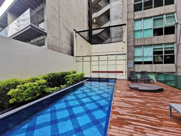 Una piscina con diseño ajedrezado, rodeada por una terraza de madera y setos verdes bajos.