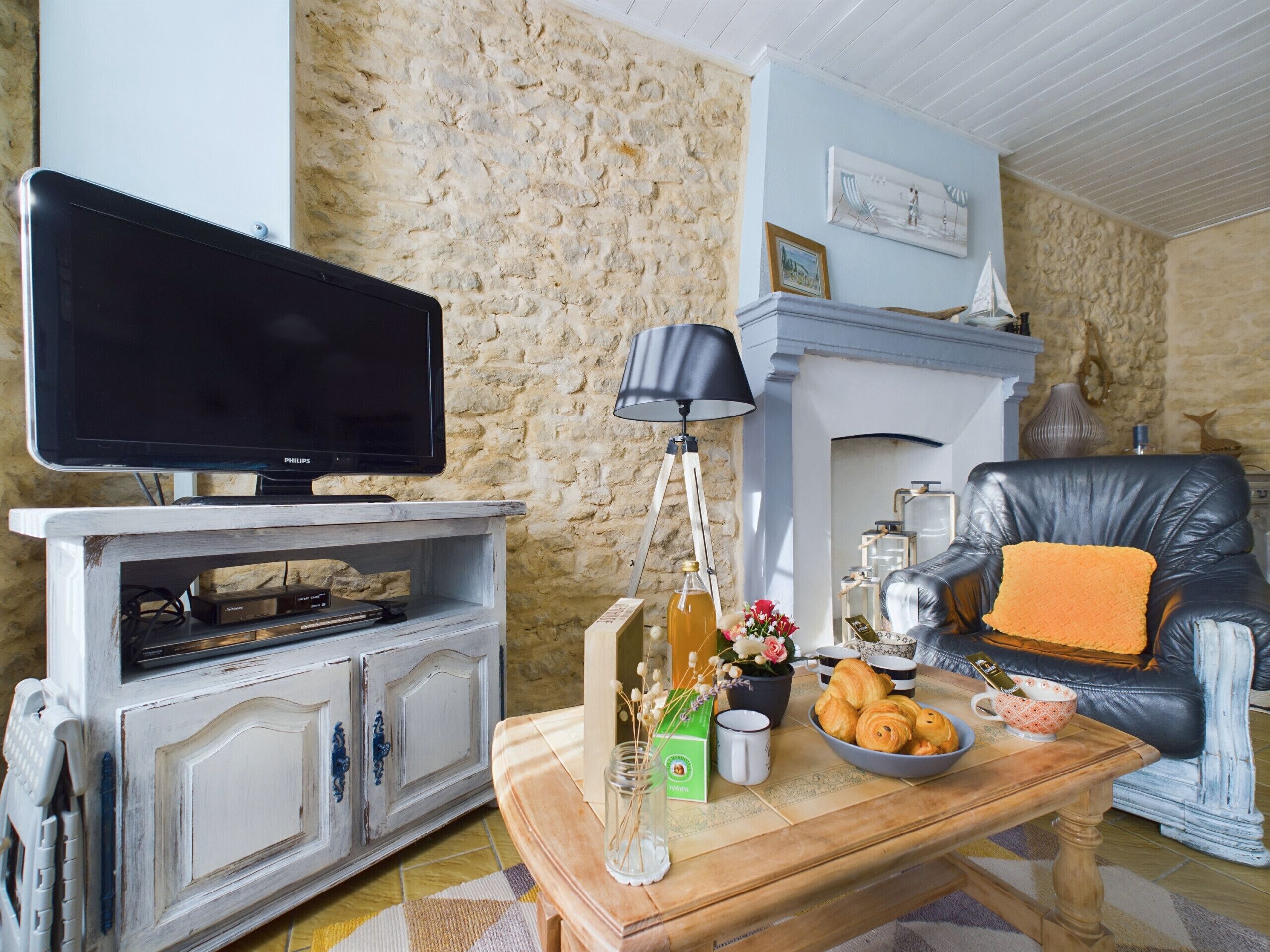 A living room with a television on a white cabinet, a fireplace, a leather armchair, and a coffee table with pastries and a vase.