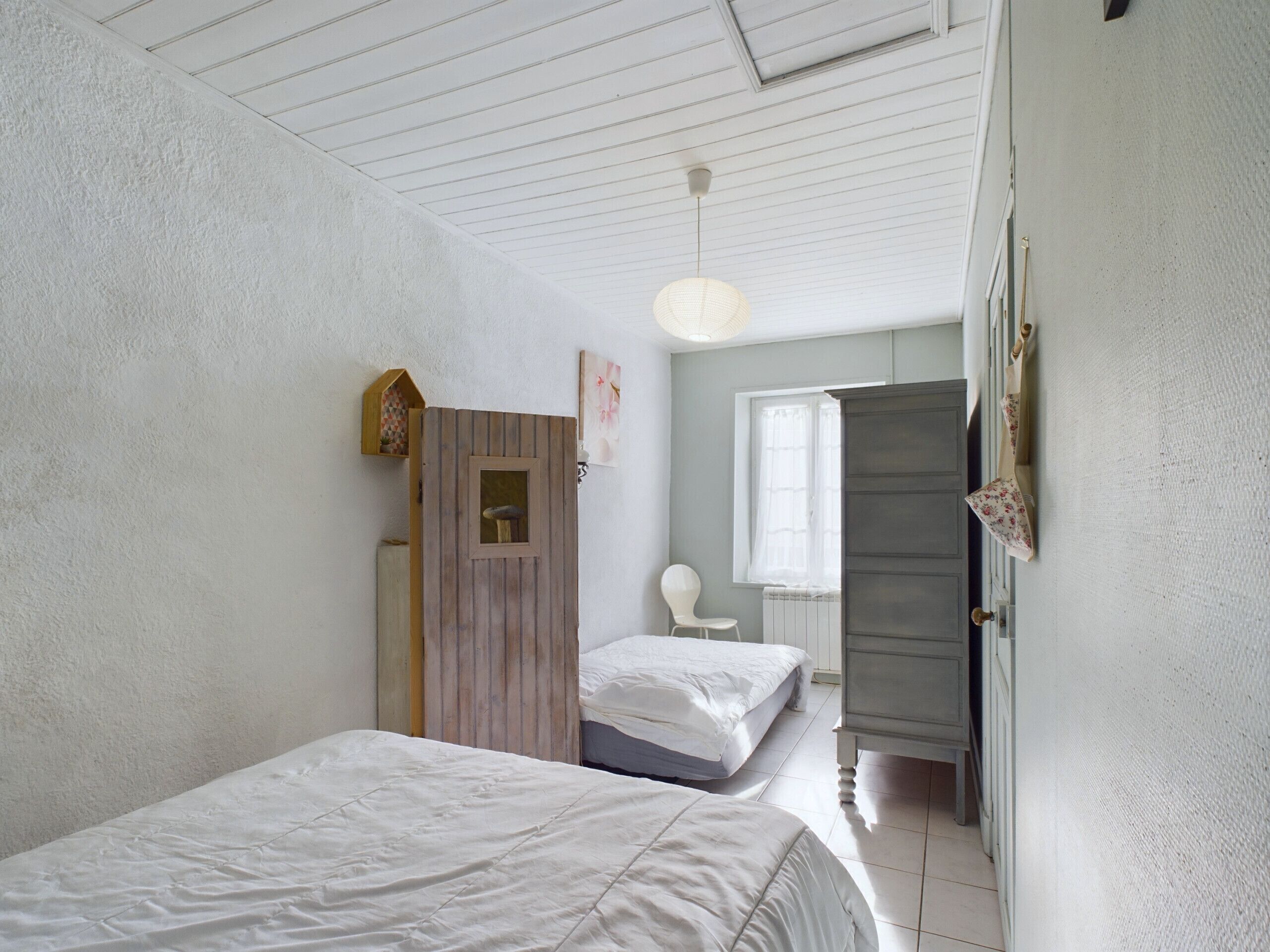 A bedroom with two beds, a wooden wardrobe, a white chair, and a window with curtains.