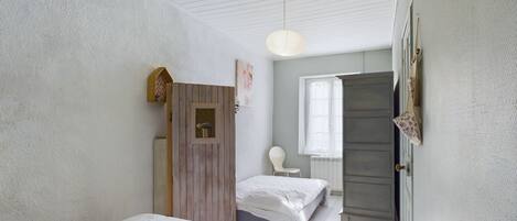 A bedroom with two beds, a wooden wardrobe, a white chair, and a window with curtains.