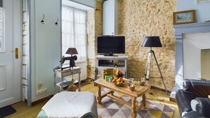 A living room with a stone wall, a television on a stand, a wooden table with a lamp, and a black leather sofa.