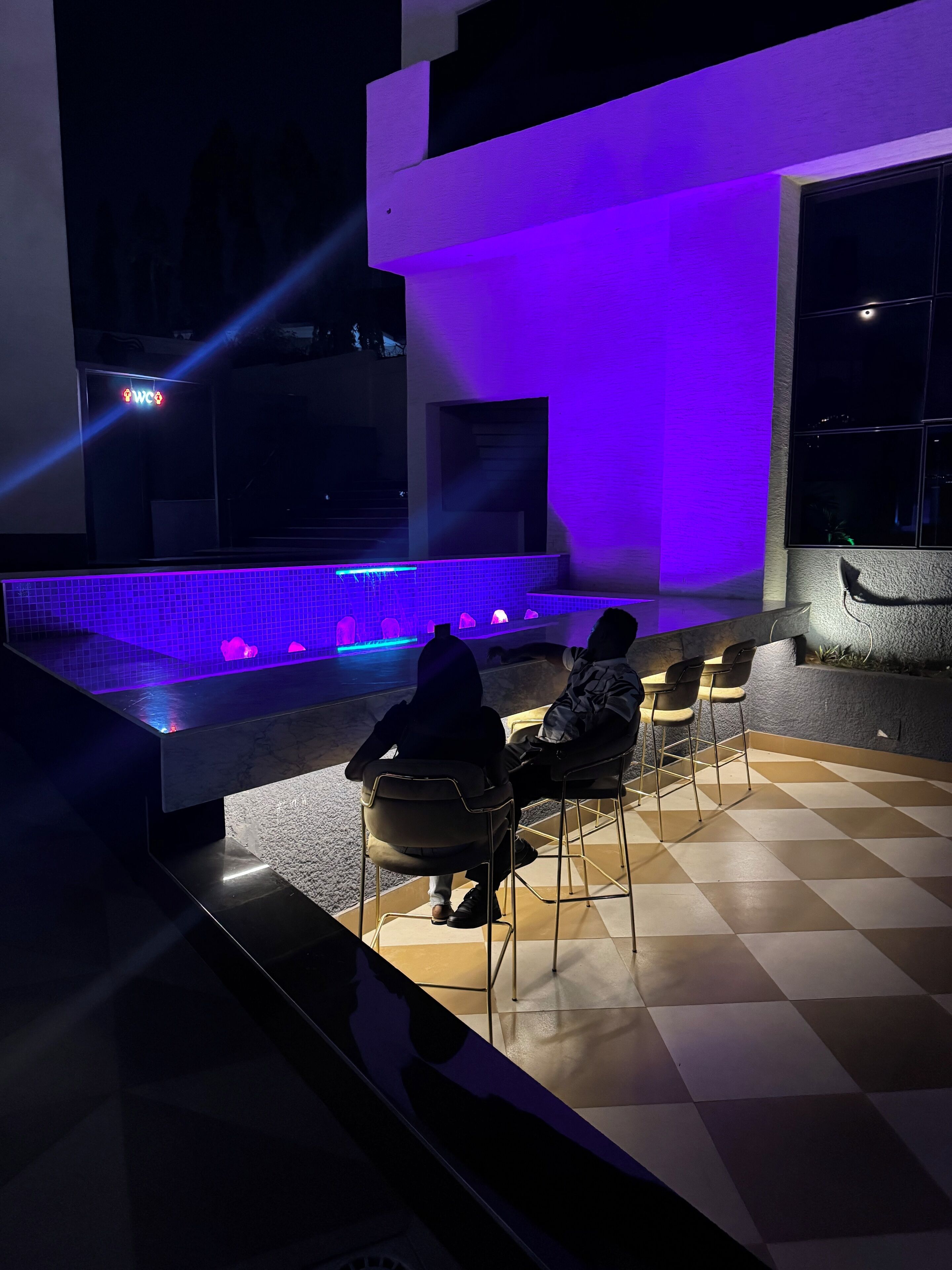 An outdoor seating area with a checkered floor, a pool with a blue-lit edge, and a modern building with a purple-lit wall.