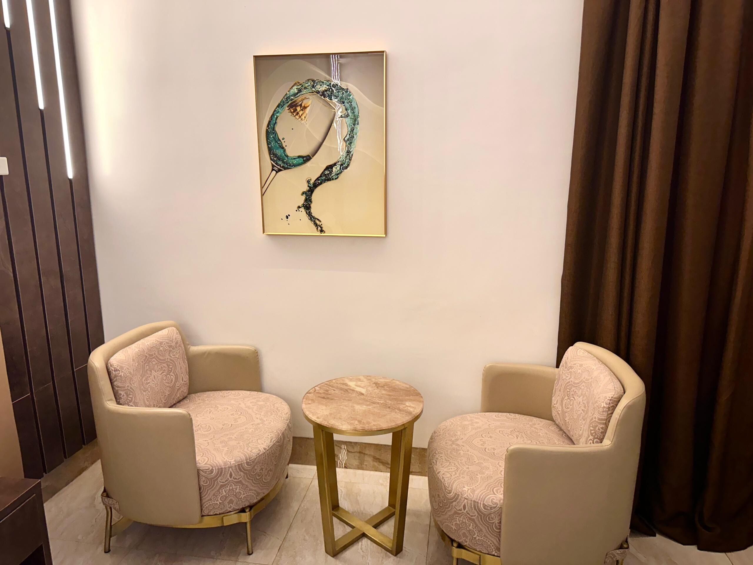 A room with two beige armchairs, a small round table, and a framed abstract painting on the wall.