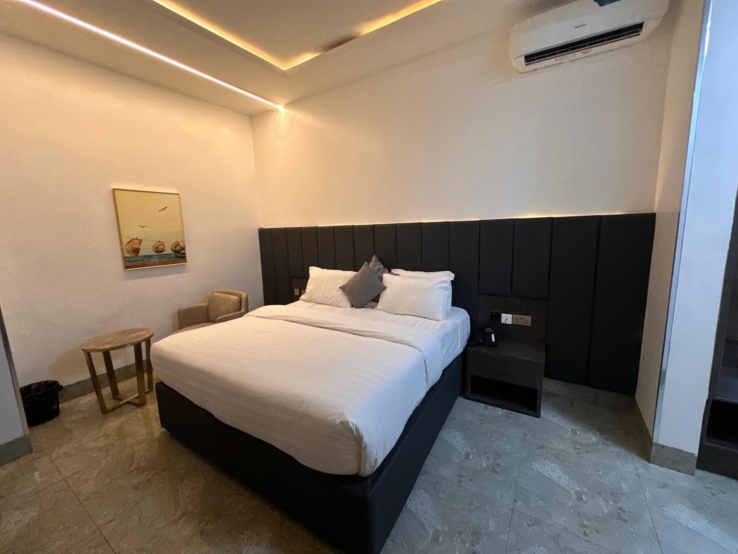 A modern hotel room with a large bed, a bedside table, a chair, and a wall-mounted air conditioner.