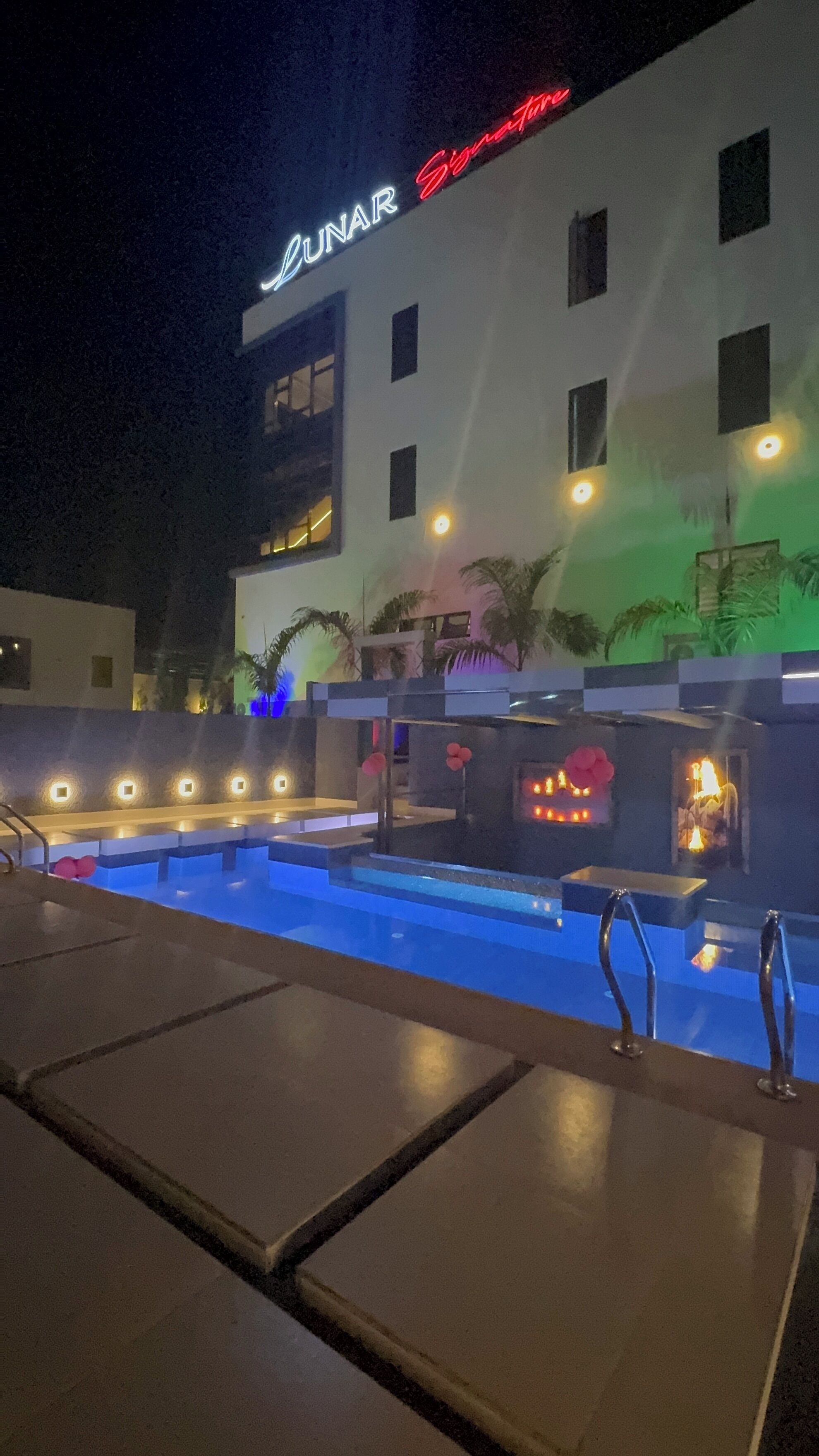 Ein Hotelpoolbereich mit einem leuchtenden Neon-Schild mit der Aufschrift "LUNAR Signature" und einem modernen Gebäude mit vielen Fenstern.