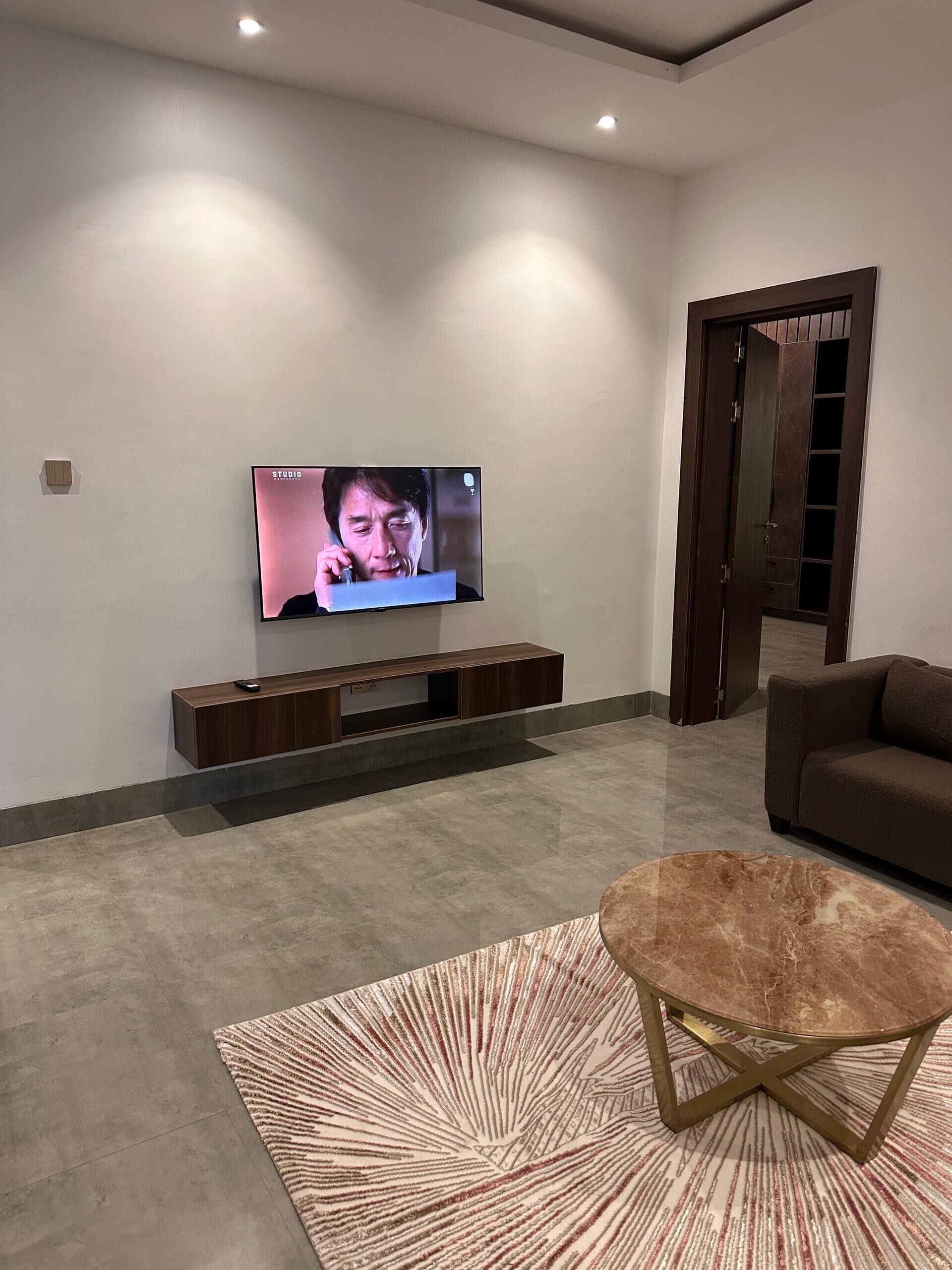 Un salon moderne avec un téléviseur à écran plat fixé au mur, un canapé brun foncé, un tapis à motifs et un meuble de rangement en bois.