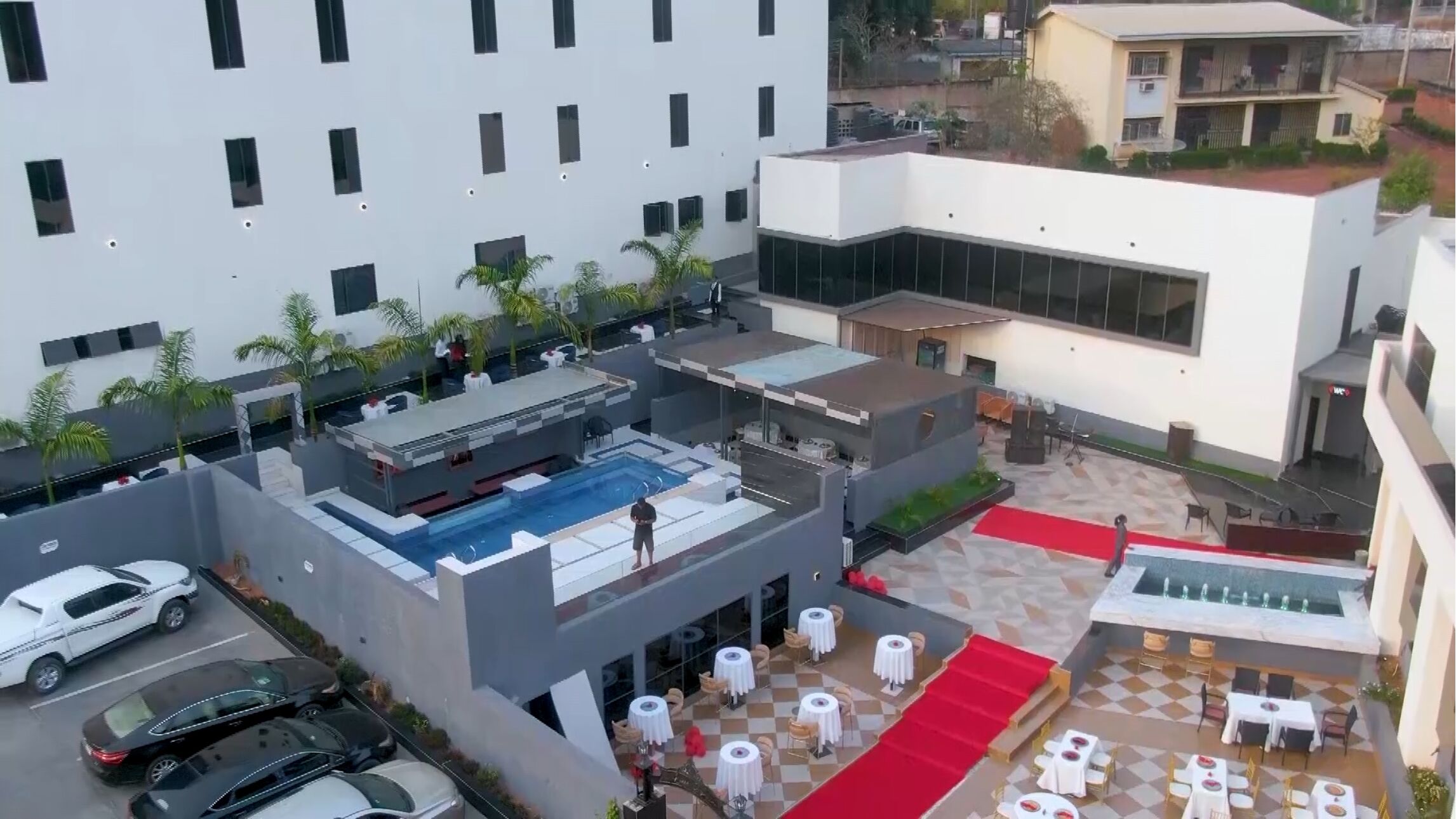 Un espace piscine aménagé sur le toit, avec plusieurs tables et chaises, entouré de palmiers et bordé d’un tapis rouge.