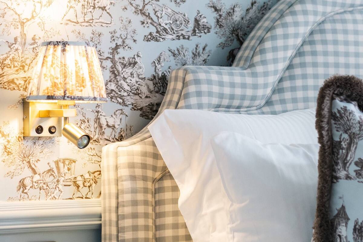 A bedroom with a checkered bedspread, a wall-mounted lamp, and a floral wallpaper.