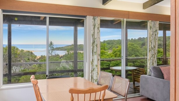 A room with a large sliding glass door leading to a balcony with a view of the ocean and greenery. There is a table with chairs and a sofa inside the room.