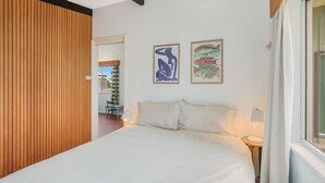 A bedroom with a bed, a wooden slat wall, a window with curtains, and two framed artworks on the wall.
