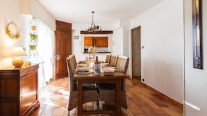 Ein Essbereich mit einem Holztisch, gedeckt für vier Personen, umgeben von Korbstühlen. Über dem Tisch hängt eine Kristallleuchter, und im Hintergrund ist eine Küche zu sehen.