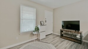 A room with a TV on a wooden stand, a door, a window with blinds, and a small table with a plant. - Cozy Central Austin Getaway, Pool, Wi-Fi, Free Parking, Near Downtown, So Co. (Austin)