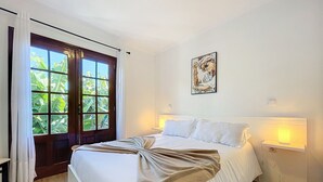 A neatly made bed with a beige throw, a large window with curtains, a framed artwork on the wall, and a bedside table with a lamp. - Sweet Home Madeira C by Atlantic Holiday (Funchal)
