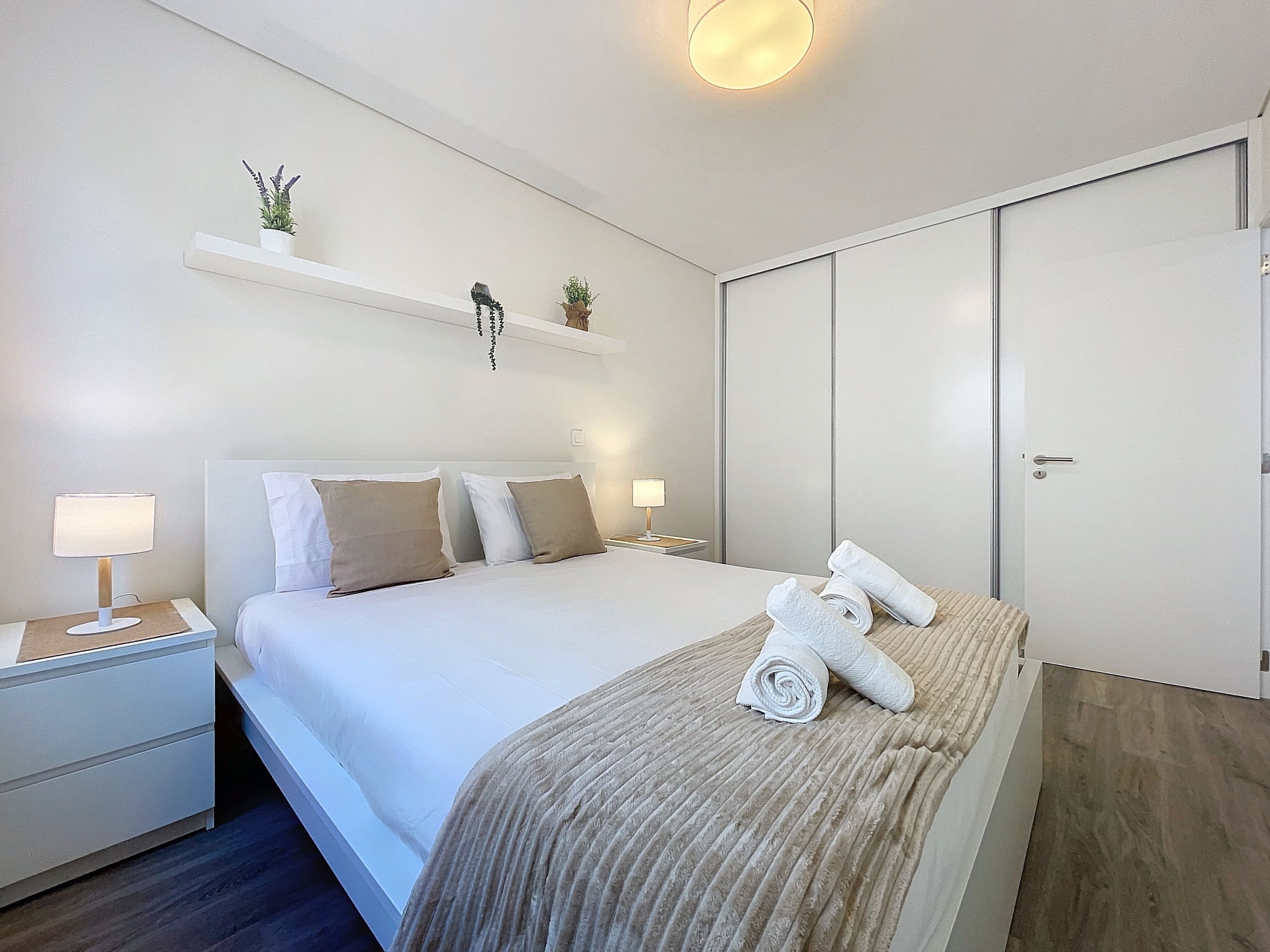 A modern hotel room with a neatly made bed, a bedside table with a lamp, a small shelf with decorative items, and a wardrobe.