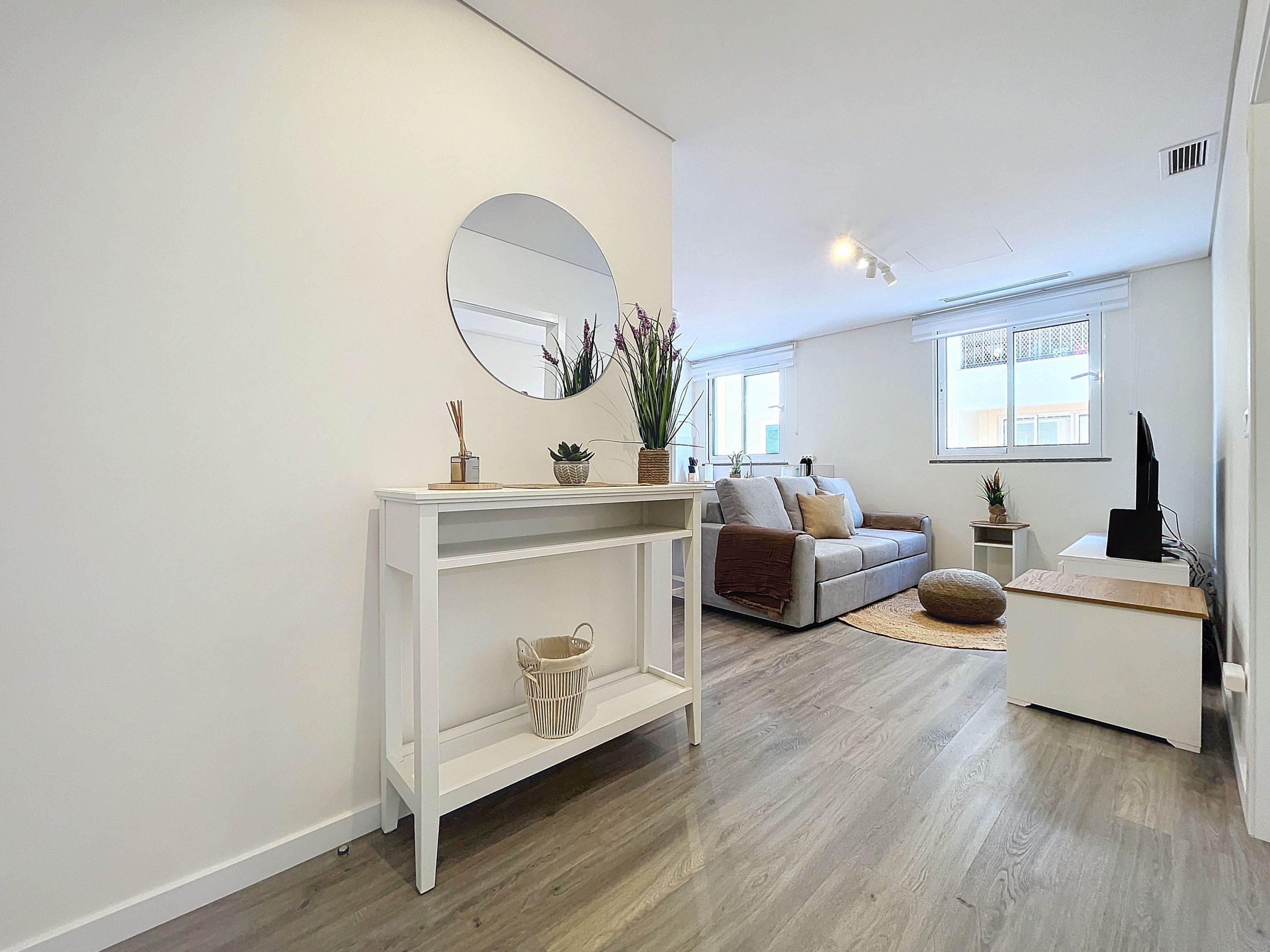 A modern living room with a white console table, a grey sofa, a round mirror, and a wooden floor.