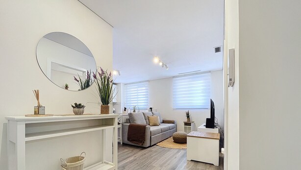 Una sala moderna con sofá, mesa de centro y televisor. Se observa una consola blanca con objetos decorativos y un espejo.