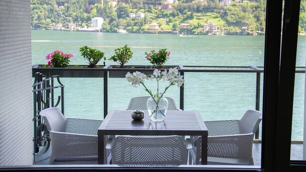 Ein Balkon mit Tisch und zwei Stühlen, mit Blick auf einen See und Häuser im Hintergrund.