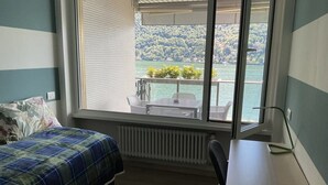 Ein Schlafzimmer mit Bett, Schreibtisch und Fenster mit Blick auf einen See und Grünflächen.