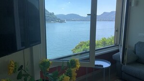 Ein Zimmer mit großem Fenster und Blick auf einen See und Berge. An der Wand ist ein Flachbildfernseher angebracht, es gibt einen kleinen runden Tisch und ein Sofa.