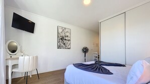 Une chambre d’hôtel moderne équipée d’un lit, d’un bureau avec une chaise, d’un téléviseur à écran plat, d’un ventilateur fixé au mur et d’une œuvre d’art encadrée.