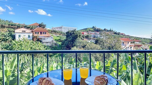 Un balcon avec deux assiettes de croissants et un verre de jus d’orange, donnant sur une colline parsemée de maisons et de verdure.