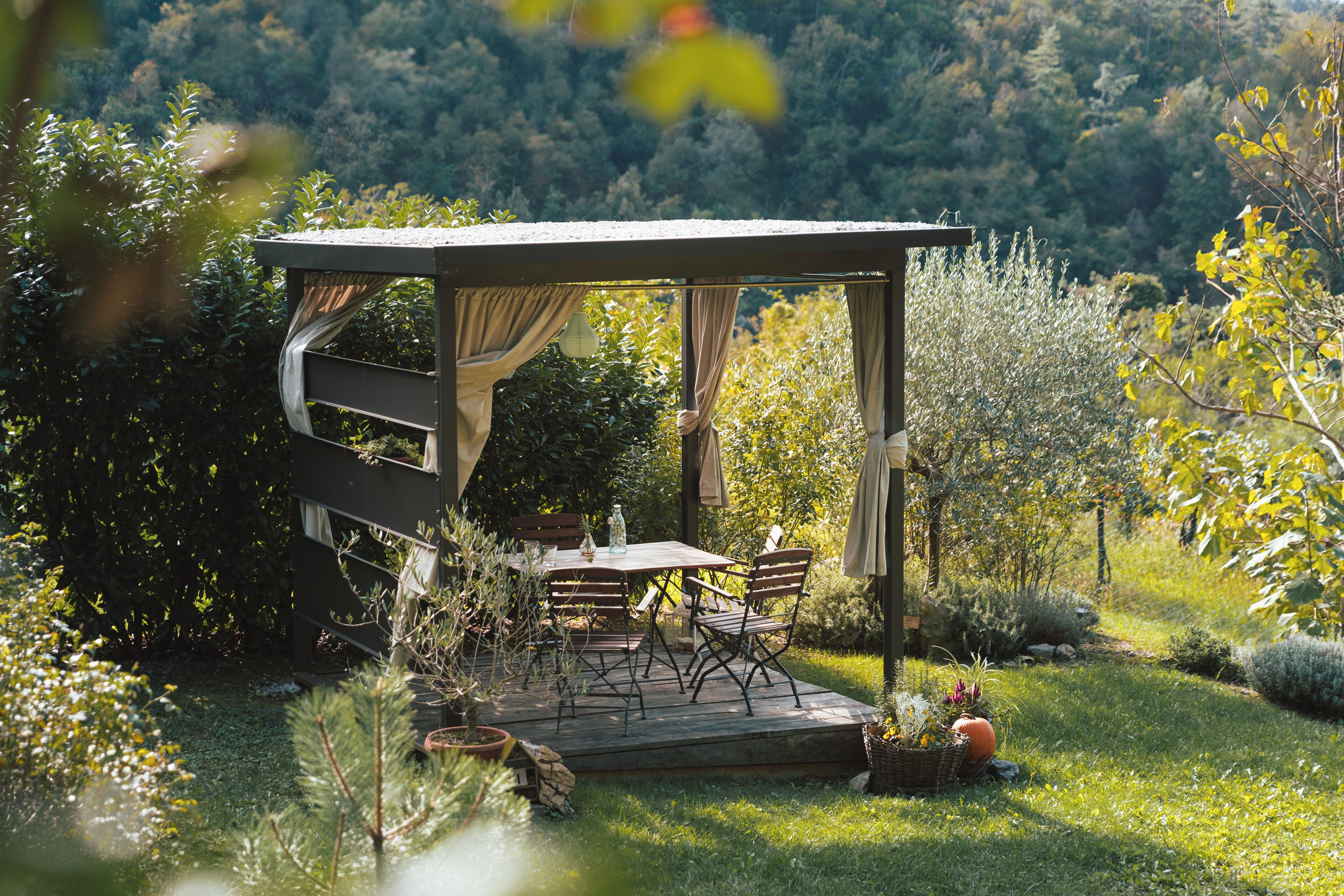 A wooden deck with a table and chairs, surrounded by lush greenery and a forested backdrop.