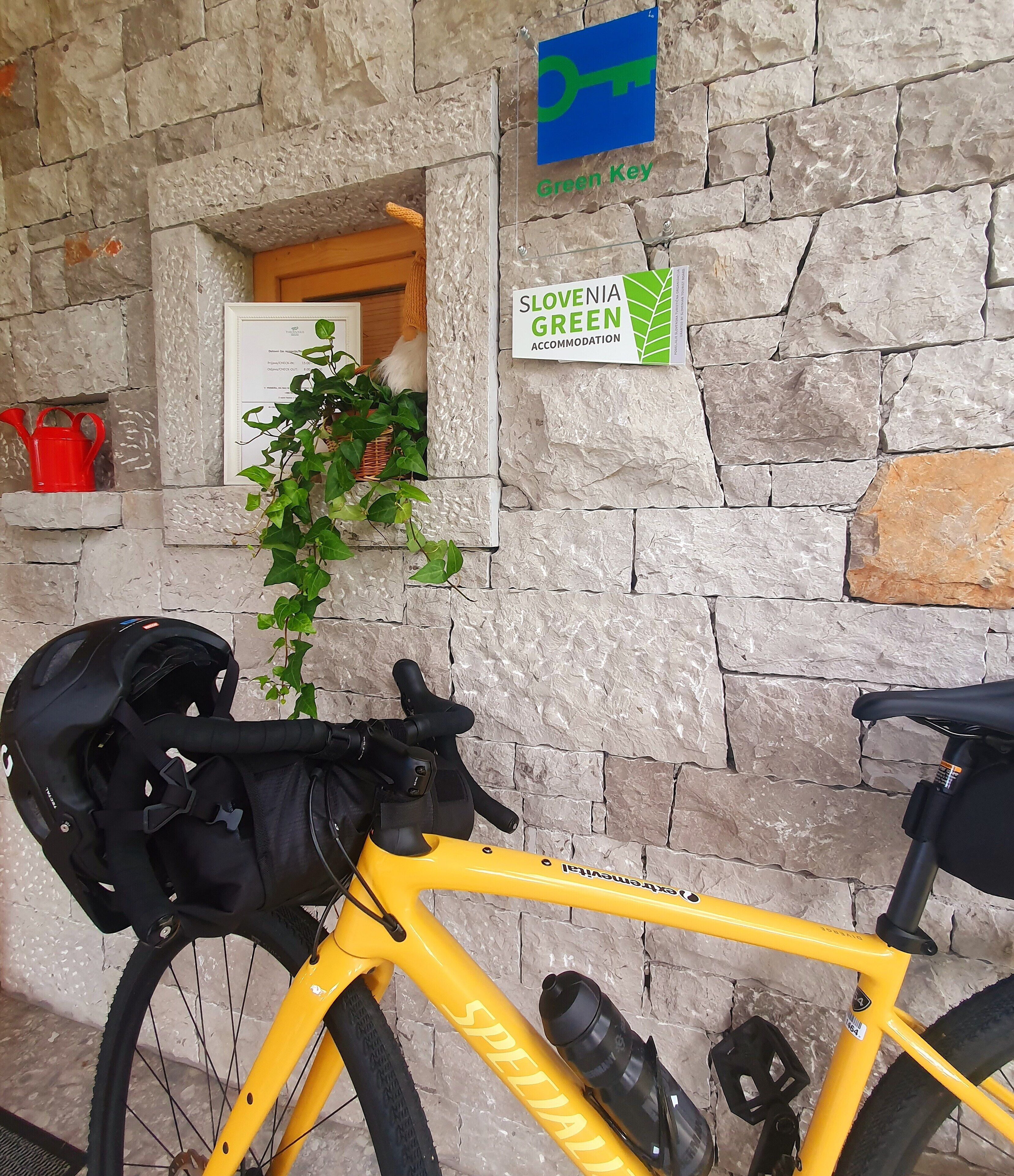 Een gele fiets met een zwarte tas, geparkeerd tegen een stenen muur met een 'Green Key' bord en een 'Slovenia Green Accommodation' bord.
