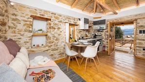 Une pièce chaleureuse avec un mur en pierre, un plafond en bois, un coin repas avec des chaises, une cuisine équipée d’une cuisinière et une vue sur la mer.
