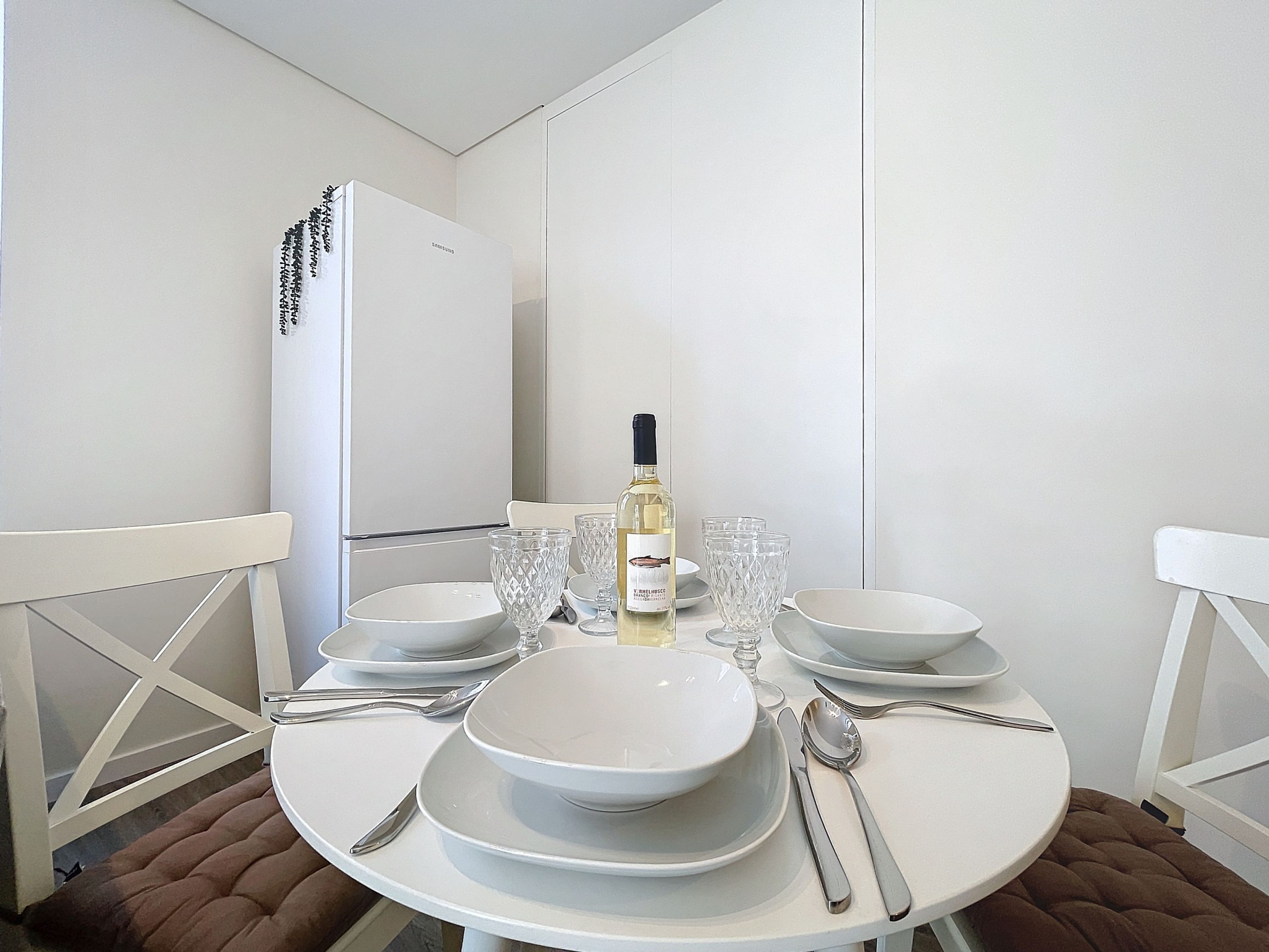 A dining table set with white plates, bowls, cutlery, and a bottle of white wine, with a white refrigerator in the background.