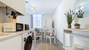 Una cocina compacta con microondas, tetera y lavadora. Cuenta con una zona de comedor con mesa y sillas, y una pequeña planta en maceta sobre un estante.