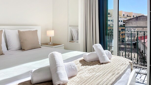 Habitación de hotel con una cama grande, una mesita de noche con lámpara, un espejo y vistas a la ciudad desde un balcón con barandilla metálica.