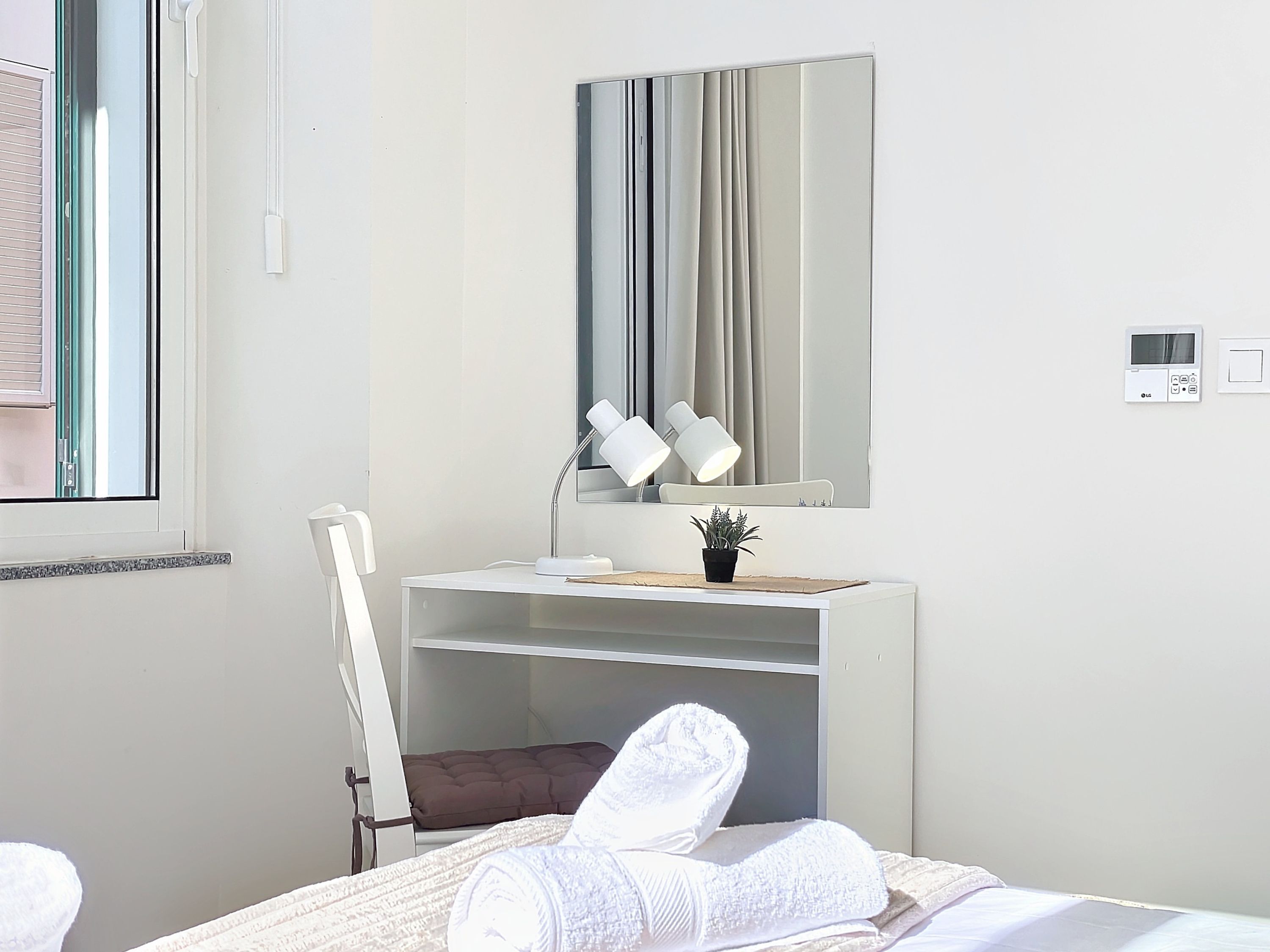 A hotel room with a bed, a desk with a mirror, a small potted plant, and a wall-mounted thermostat.
