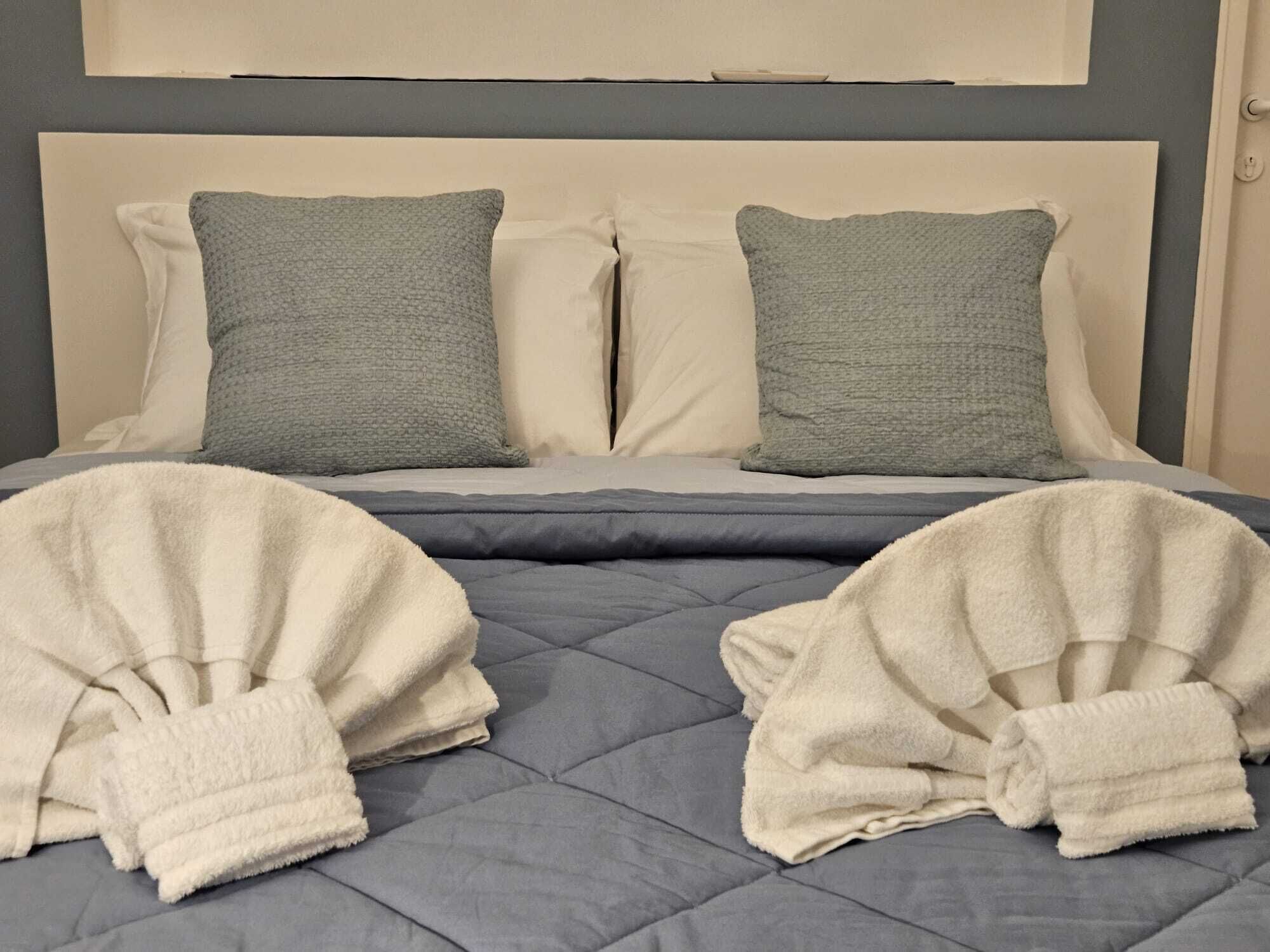 A bed with a grey quilt, two grey pillows, and two white towels.