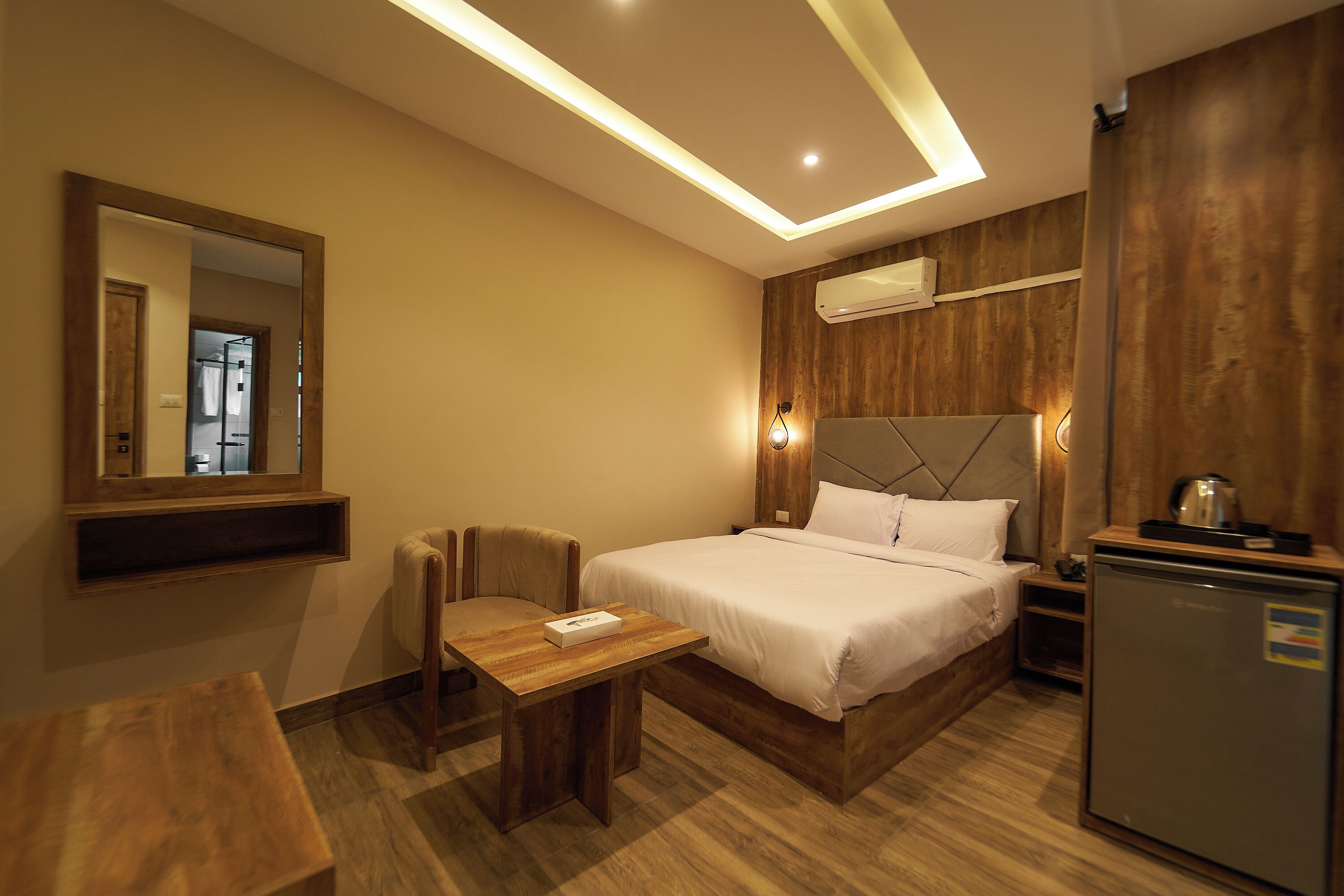 A hotel room with a bed, a desk, a chair, a mirror, a small table, a refrigerator, and an air conditioning unit.