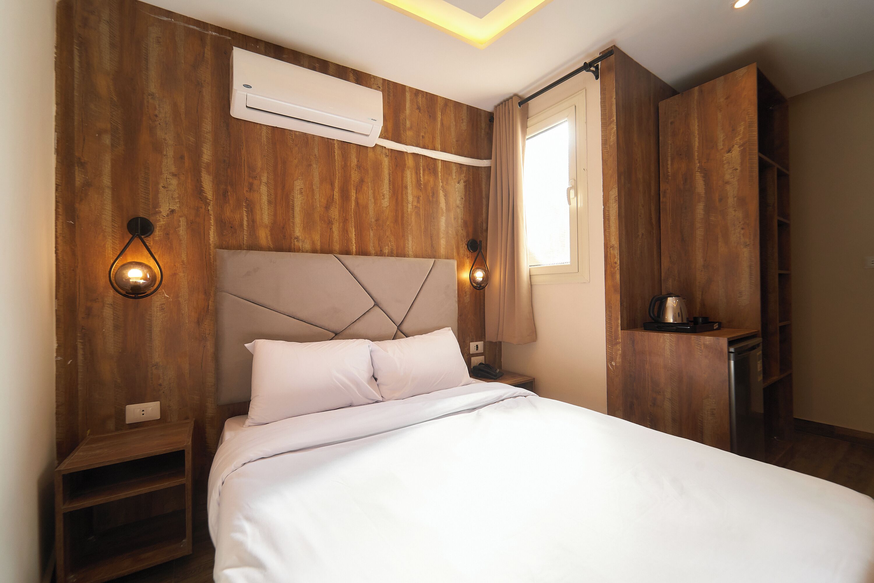 A bedroom with a wooden headboard, a bed with white linens, a nightstand with a lamp, and a built-in kitchen area.