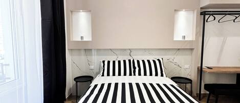 A modern bedroom with a black and white striped bed, two bedside tables with lamps, a wooden desk, and a large window with sheer curtains.
