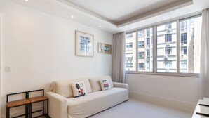 A modern living room with a white sofa, a small wooden bench, and a large window offering a view of buildings.