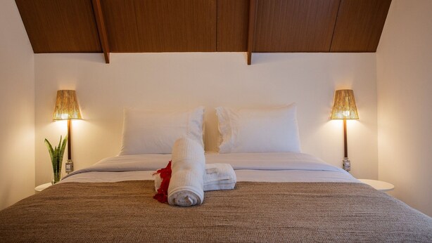 A neatly made bed with white linens and a rolled towel, two bedside lamps, and a plant on a bedside table. - Cabana Mulungu (Fernando de Noronha)