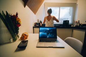 Eine Küche mit einem Laptop auf dem Tisch, einer Blumenvase und einem Fenster mit Blick auf Bäume.