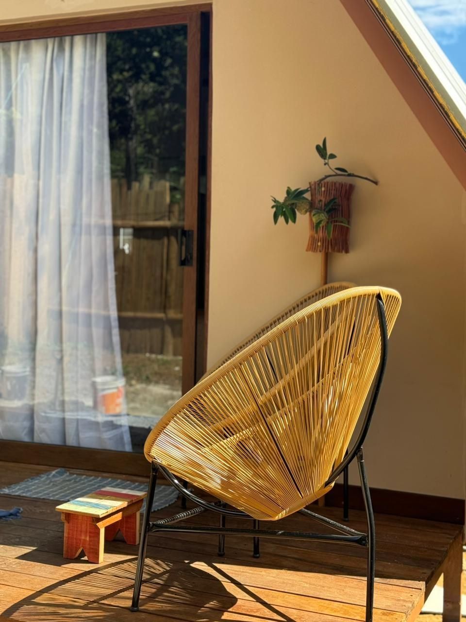 A wicker chair with a metal frame on a wooden deck, a small stool, and a hanging plant.