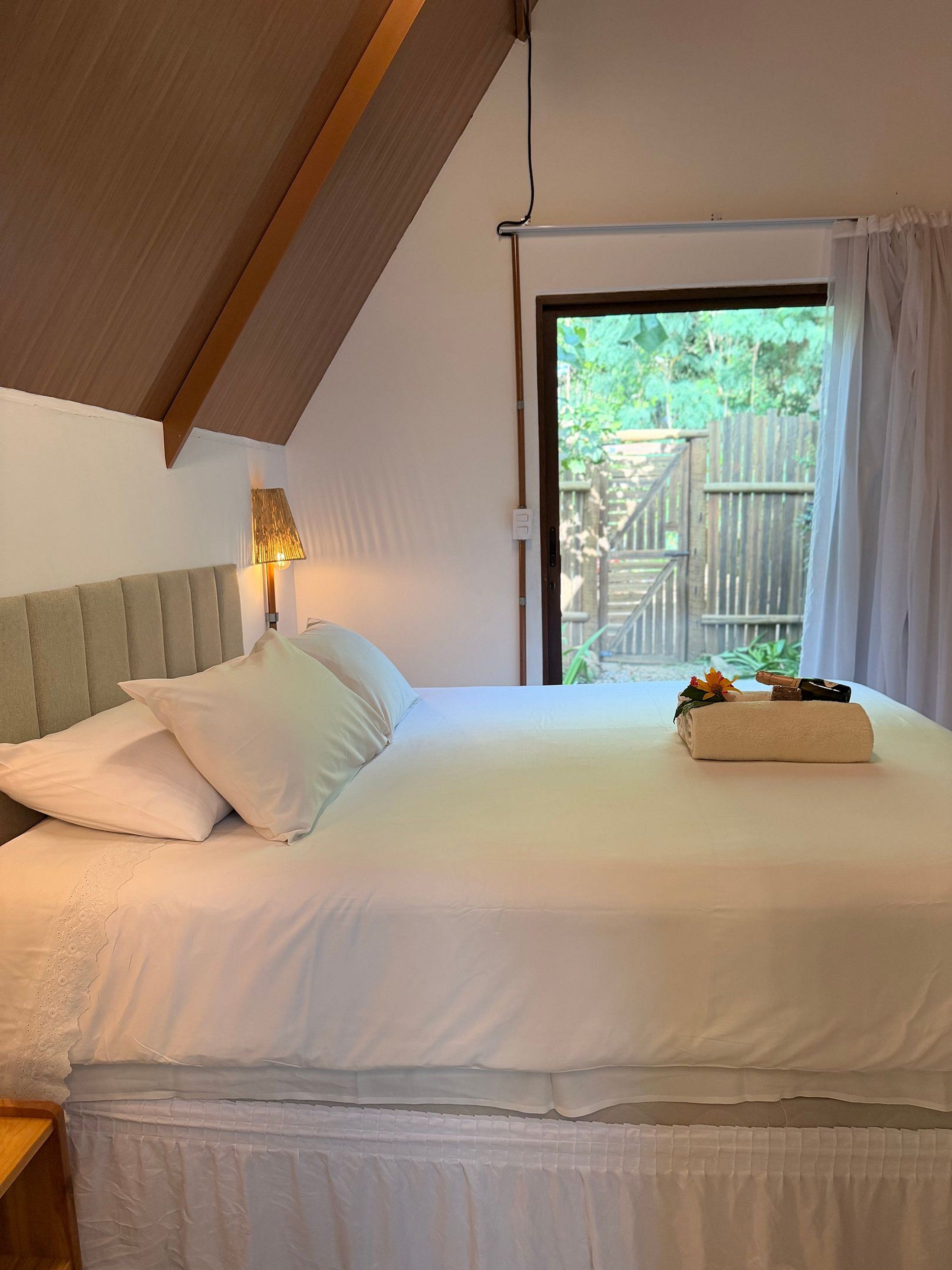 A neatly made bed with pillows and a folded towel, a bedside lamp, and a view of greenery outside.
