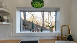 A kitchen with a window view of a forest, a white countertop, a dishwasher, and a gold faucet. - Woodland Nook (Alpine)