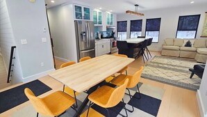 Eine moderne Küche mit integriertem Kühlschrank, ein Essbereich mit Holztisch und gelben Stühlen sowie ein Wohnzimmer mit beiger Couch und großem Fenster.