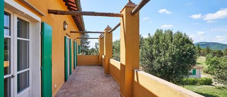 Un balcón estrecho con suelo de baldosas, puertas verdes y vistas a vegetación y colinas al fondo.